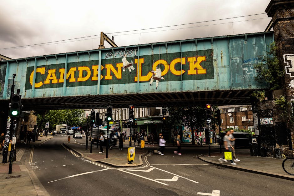 A large, weathered railway bridge with a painted sign reading 'CAMDEN LOCK' in bold yellow letters, spanning over a busy urban street. The sign features an illustration of two children hanging from th