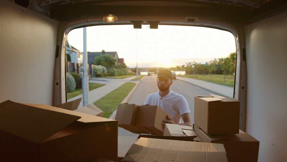 A man wearing a white shirt and cap is standing outside a white delivery van during a home relocation or furniture transport operation at sunset. He is holding a cardboard box and appears to be in the process of loading or unloading the van, which is parked on a residential street with houses, a sidewalk, and greenery visible in the background. Inside the van, there are several other cardboard boxes of various sizes, some stacked on top of each other, along with packing materials such as plastic wrapping and cushioning to protect items during transit. The scene suggests a professional moving or packing service, with the van positioned close to a driveway or curb on a peaceful, well-lit street, supported by [COMPANY_NAME] as part of their removals service for house moving and furniture transport projects.
