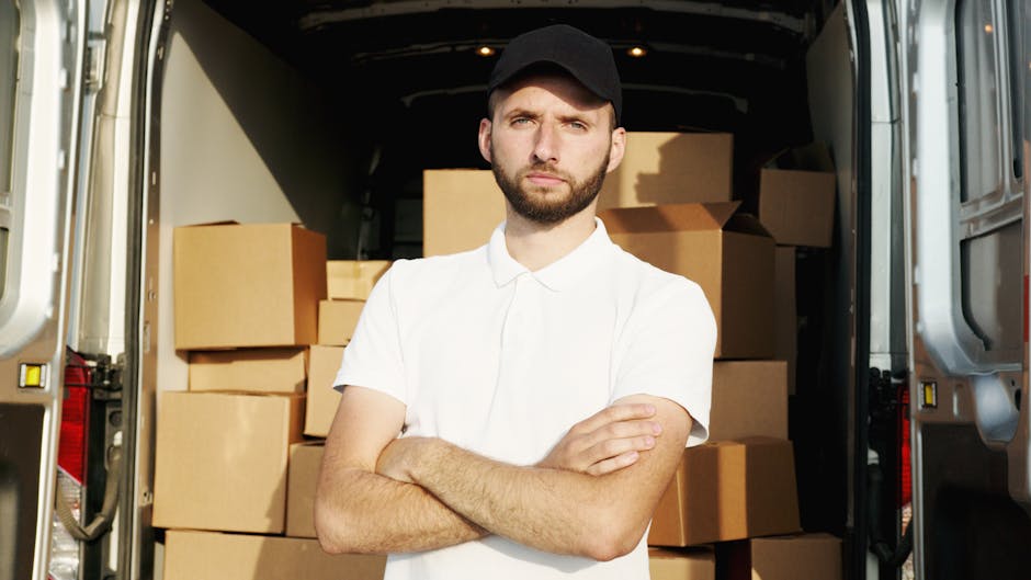 A man with a short beard, wearing a black cap and a white short-sleeved shirt, stands with arms crossed inside the back of a delivery van, which is filled with neatly stacked cardboard boxes. The van's interior is lit with natural daylight, highlighting the plain brown boxes arranged on the floor and sides. The van's rear doors are open, revealing the surrounding environment, possibly on a street or driveway. The boxes are of various sizes, some wrapped in plastic or secured with packing tape, indicating a home relocation or furniture transport process. Brixton Man and Van's vehicle is positioned on pavement, ready for loading or unloading, supporting professional removals and moving services for household or trade clients.
