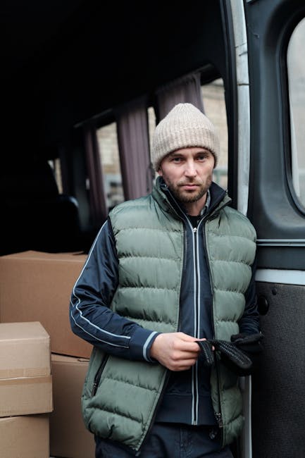 A man wearing a white shirt and cap is standing outside a white delivery van during a home relocation or furniture transport operation at sunset. He is holding a cardboard box and appears to be in the process of loading or unloading the van, which is parked on a residential street with houses, a sidewalk, and greenery visible in the background. Inside the van, there are several other cardboard boxes of various sizes, some stacked on top of each other, along with packing materials such as plastic wrapping and cushioning to protect items during transit. The scene suggests a professional moving or packing service, with the van positioned close to a driveway or curb on a peaceful, well-lit street, supported by [COMPANY_NAME] as part of their removals service for house moving and furniture transport projects.