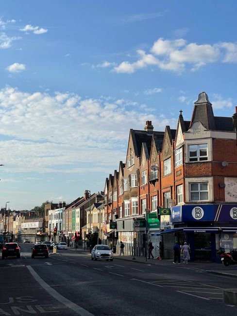 A street scene in Brixton showing a row of multi-storey residential buildings with brick facades and bay windows, situated along a busy pavement. Several cars are seen driving along the road, while pedestrians walk on the sidewalk, some near a shopfront or bus stop. The sky is partly cloudy with patches of blue, and the sunlight provides natural lighting to the scene. This urban environment reflects the typical setting for home relocations and moving logistics, with commercial storefronts visible at street level. Brixton Man and Van is aware of such surroundings when providing removals services, including packing, loading, and furniture transport for local house moves within the area.