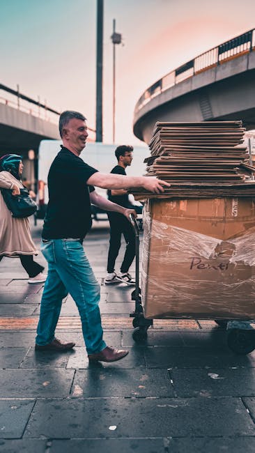 A man in casual clothing, including a black top and blue jeans, is pushing a trolley loaded with large cardboard boxes wrapped in plastic and packing materials. The trolley is positioned on a paved outdoor area beneath a bridge or overpass structure, with tall lamp posts and railings visible in the background. Several other individuals, some carrying smaller boxes or bags, are nearby, indicating a scene of home relocation or furniture transport during a moving process. The image captures a moment of loading or unloading, with the presence of Brixton Man and Van's removals team, highlighting a professional moving service involved in packing and transportation logistics for house removals within the Brixton area.